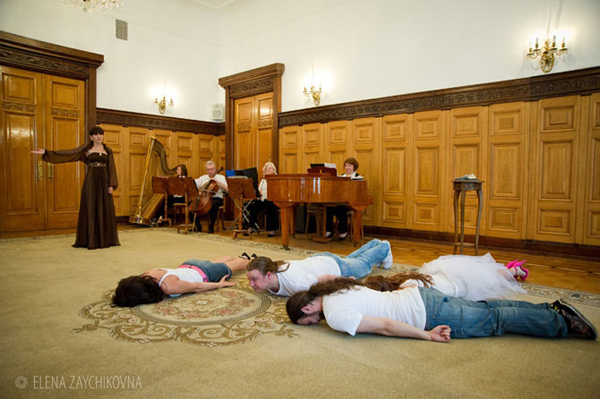 People planking on a rug while classical musicians play in a grand hall