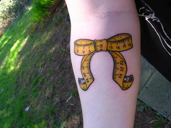 Forearm tattoo of a yellow measuring tape tied into a bow