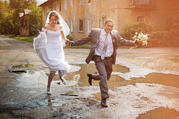 Bride and groom leaping through street puddles hand in hand