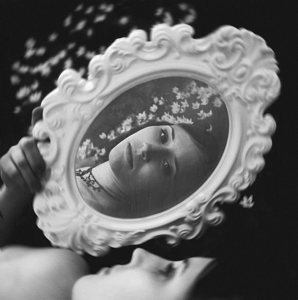 Woman holding an ornate oval mirror above her face with petal-like shapes in the air